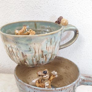 May include: Two handmade ceramic mugs stacked on top of each other. The mugs are light blue with brown accents and feature small, whimsical animal figurines inside and on the handles. The mugs have a rustic, textured appearance.