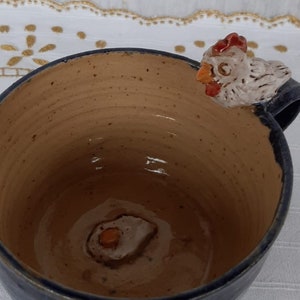 Può includere: Una tazza in ceramica blu con interno marrone e una piccola statuetta di pollo sul manico. La tazza ha una glassa macchiettata e un design a forma di uovo marrone all'interno.