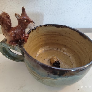 Könnte beinhalten: Eine Keramiktasse mit einer Eichhörnchenfigur an der Seite. Die Tasse hat einen grünen Henkel und eine braune, gesprenkelte Glasur. Das Innere der Tasse ist beige.