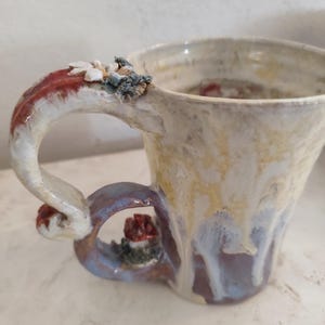 Op de afbeelding: Een keramische mok met een unieke, gebeeldhouwde handgreep met een bloemenmotief. De mok is geglazuurd in wit en geel met een vleugje paars en rood.