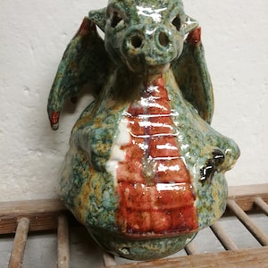 Könnte beinhalten: Eine Keramik-Drachenfigur mit grüner, brauner und roter Glasur. Der Drache hat ein schwarzes Horn und große Flügel.