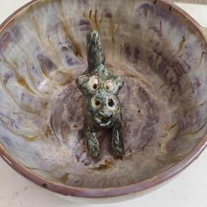 May include: A ceramic bowl with a mottled purple, brown, and cream glaze featuring a small, sculpted green creature with two eyes and a tail positioned in the center of the basin. The bowl has a smooth, rounded rim and a deep, circular interior.