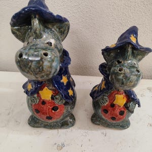 Könnte beinhalten: Zwei Keramik-Drachenfiguren. Die Drachen sind blau und grün mit einem Zaubererhut und Umhang. Jeder Drache hat ein rotes Marienkäfer-Design auf dem Bauch mit gelben Sternen. Die Figuren stehen auf einer weißen Oberfläche.