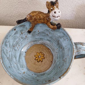 Könnte beinhalten: Eine blaue Keramiktasse mit einem braunen und weißen Pferdefiguren-Griff. Die Tasse hat ein braunes Inneres mit einem gelben Blumendesign.