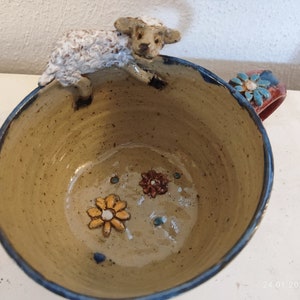 Può includere: Una tazza in ceramica con un bordo blu e un manico marrone. La tazza è decorata con una statuetta di pecora in ceramica bianca e fiori colorati.