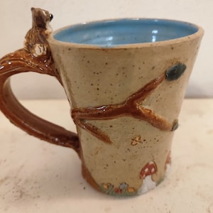 Könnte beinhalten: Eine handgefertigte Keramik-Tasse mit hellbraunem Äußeren und blauem Innenraum. Der Griff ist ein brauner Ast mit einem Eichhörnchen darauf. Die Tasse ist mit bemalten Pilzen, Blättern und einem Schmetterling verziert.
