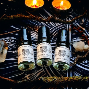 Può includere: Tre bottiglie di profumo Cherry Bear con tappi neri, ciascuna con un'etichetta con un'illustrazione di un orso. Le bottiglie sono su una superficie scura a motivi, con candele accese e cristalli nelle vicinanze. Il testo "WOLF BERRY RITUALS" è sull'etichetta.