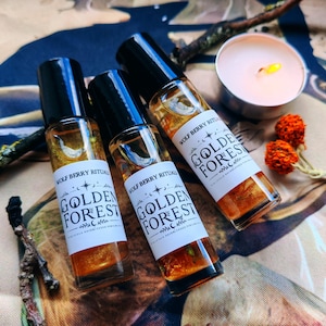 Op de afbeelding: Drie doorzichtige glazen rollerball parfumflesjes met zwarte doppen, elk voorzien van een wit etiket met de tekst "WOLF BERRY RITUALS" en "GOLDEN FOREST" in zwarte inkt. De flesjes bevatten een goudkleurige vloeistof met zichtbare botanische elementen.