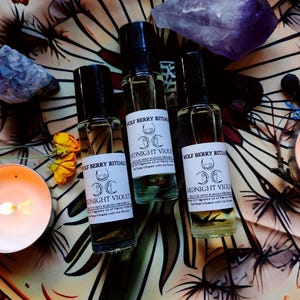 Könnte beinhalten: Drei Glasflaschen "Wolf Berry Ritual" Duftöl mit schwarzen Kappen und weißen Etiketten mit der Aufschrift "Midnight Violet". Die Flaschen stehen auf einem gemusterten Tuch mit Kristallen, Blumen und einer brennenden Kerze.