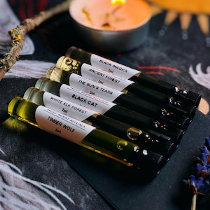 Puede incluir: Una colección de pequeños viales de vidrio llenos de líquidos de colores, cada uno etiquetado con un nombre único como "Black Magick" y "Timber Wolf". Los viales están dispuestos sobre una superficie oscura, con una vela encendida y elementos decorativos en el fondo.