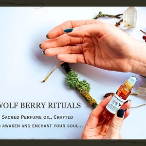 May include: A small, amber-colored perfume oil bottle with a rollerball applicator is held in a hand with dark blue nail polish. The bottle is labeled "Wolf Berry Rituals." The background includes twigs, moss, and text that reads "Sacred Perfume Oil, Crafted to Awaken and Enchant Your Soul..."