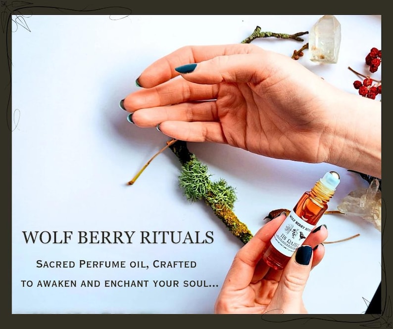 May include: A small, amber-colored perfume oil bottle with a rollerball applicator is held in a hand with dark blue nail polish. The bottle is labeled "Wolf Berry Rituals." The background includes twigs, moss, and text that reads "Sacred Perfume Oil, Crafted to Awaken and Enchant Your Soul..."