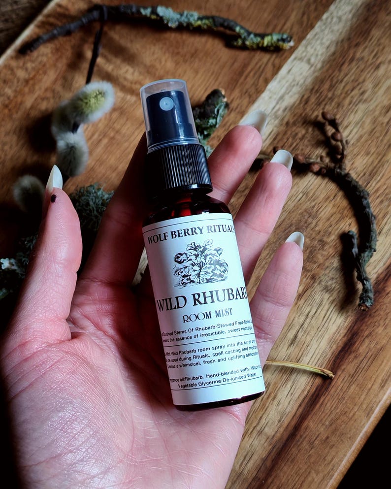 May include: A dark amber glass spray bottle of "Wild Rhubarb Room Mist" held in a hand. The label features text and a botanical illustration. The bottle has a black spray nozzle. The background is a wooden surface with dried plants.