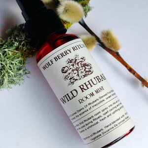 May include: A red glass bottle of "WILD RHUBARB ROOM MIST" with a black spray nozzle. The label features text and a floral illustration. The bottle is positioned near sprigs of pussy willow and moss.