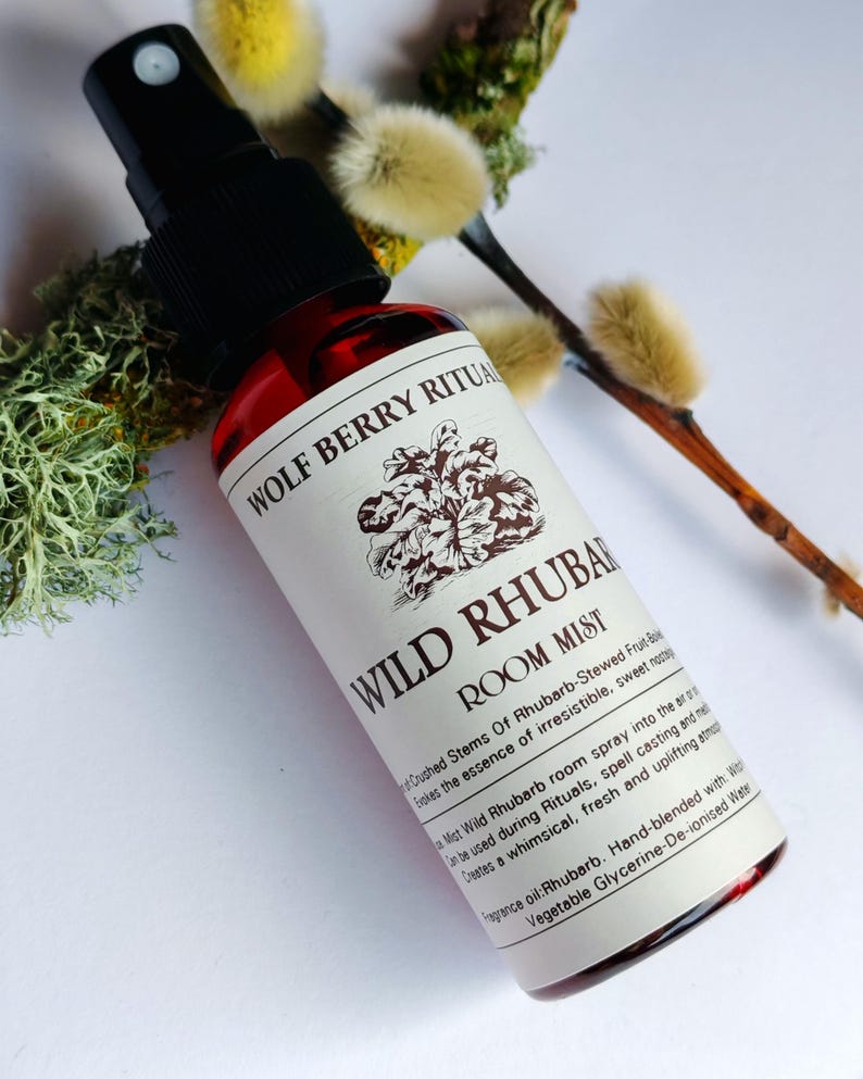 May include: A red glass bottle of "WILD RHUBARB ROOM MIST" with a black spray nozzle. The label features text and a floral illustration. The bottle is positioned near sprigs of pussy willow and moss.