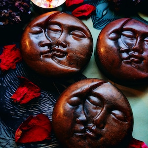 Pode incluir: Três esculturas marrons em forma de lua com rostos, dispostas sobre uma superfície estampada com pétalas vermelhas. Uma vela acesa e cristais de ametista também estão presentes. Tema celestial.