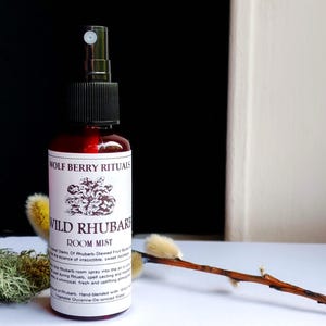 May include: A dark red glass bottle of "Wild Rhubarb Room Mist" by Wolf Berry Rituals. The label features a floral illustration and product information. The bottle has a black spray nozzle. The image includes a sprig of pussy willow.