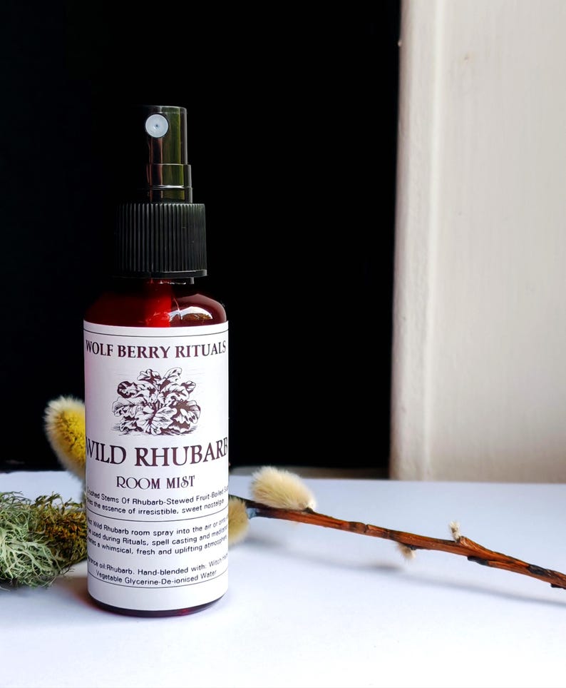May include: A dark red glass bottle of "Wild Rhubarb Room Mist" by Wolf Berry Rituals. The label features a floral illustration and product information. The bottle has a black spray nozzle. The image includes a sprig of pussy willow.