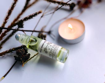 May include: A clear glass roller bottle with a silver cap and a white label that reads "Wolf Berry Ritual Ancient Forest" with a drawing of a tree and a crescent moon. The bottle is sitting on a white surface with dried branches and a lit candle.
