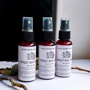 May include: Three red glass spray bottles with black caps and clear spray nozzles. Each bottle has a white label with the text "WOLF BERRY RITUALS WILD RHUBARB ROOM MIST" and a floral illustration. The bottles are arranged in a row.