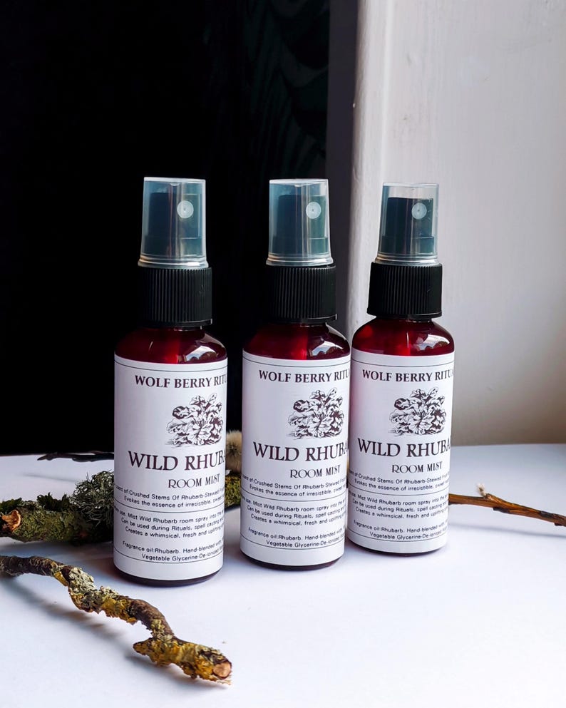 May include: Three red glass spray bottles with black caps and clear spray nozzles. Each bottle has a white label with the text "WOLF BERRY RITUALS WILD RHUBARB ROOM MIST" and a floral illustration. The bottles are arranged in a row.