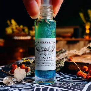 Könnte beinhalten: Eine kleine, transparente Glas-Roll-on-Flasche, gefüllt mit einer geschichteten, schimmernden blauen und grünen Flüssigkeit. Das Etikett trägt die Aufschrift "WOLF BERRY RITUALS" und "DANCING MOTH" mit einer Mottenillustration. Die Flasche wird von Fingern gehalten.