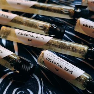 Puede incluir: Primer plano de varios pequeños viales de vidrio llenos de un líquido dorado y brillante. Cada vial tiene una tapa negra y una etiqueta blanca con el texto "CELESTIAL RAIN" y el volumen, 1 ml o 2 ml.