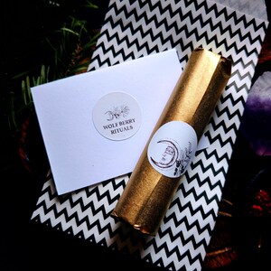 May include: A gold-wrapped item with a white sticker featuring a crescent moon and face design, next to a white card with the text "WOLF BERRY RITUALS" and a black and white zigzag patterned paper. The items are arranged on a dark surface.