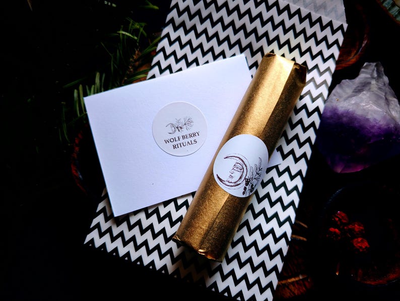 May include: A gold-wrapped item with a white sticker featuring a crescent moon and face design, next to a white card with the text "WOLF BERRY RITUALS" and a black and white zigzag patterned paper. The items are arranged on a dark surface.