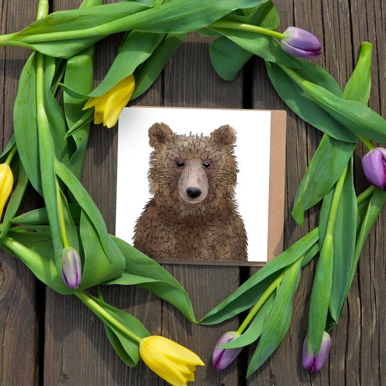 May include: A greeting card with a brown bear cub illustration on a white background. The card is surrounded by purple and yellow tulips.