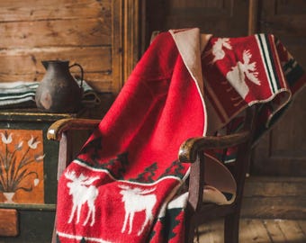 May include: A red throw blanket featuring a repeating pattern of white moose silhouettes and dark green pine trees. The blanket is draped over a wooden rocking chair, displaying a cream-colored underside and a finished edge with visible stitching.