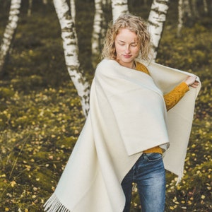 May include: A woman wearing a white shawl with a fringe, a yellow sweater, and blue jeans stands in a birch tree forest. The ground is covered in fallen leaves.