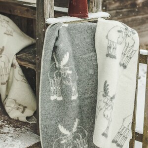 May include: Two wool blankets with a moose pattern. One blanket is white with grey moose and the other is grey with white moose. The blankets are hanging on a wooden porch railing with a red teapot on top.