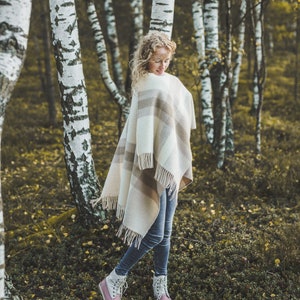 May include: A woman wearing a white and brown plaid blanket scarf stands in a birch tree forest. She is wearing blue jeans and white and pink hiking boots.
