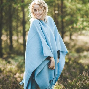 May include: A woman wearing a light blue poncho with a fringe trim, standing in a field of purple wildflowers.