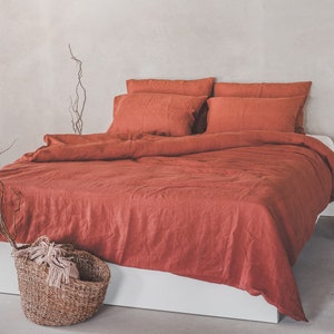May include: A rust-colored linen duvet cover and pillowcases on a white bed frame. The bedding is wrinkled and has a natural, relaxed look. There is a woven basket with a white throw blanket in the foreground.