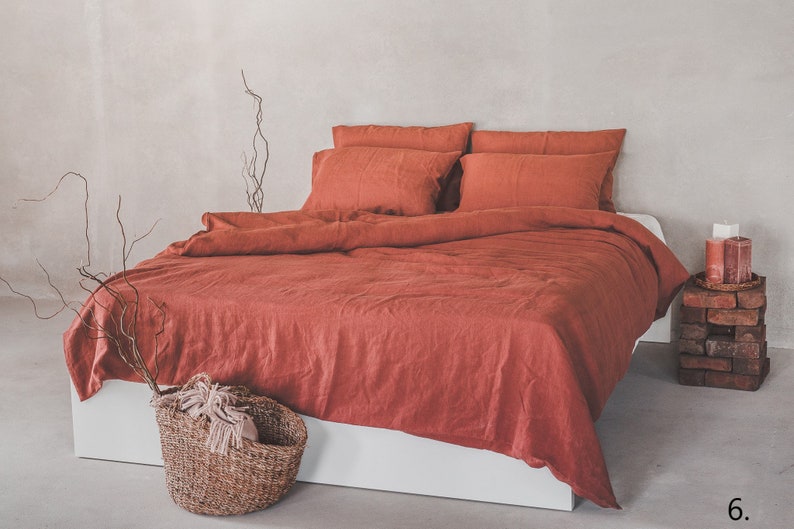 May include: A rust-colored linen duvet cover and pillowcases on a white bed frame. The bedding is wrinkled and has a natural, relaxed look. There is a woven basket with a white throw blanket in the foreground.