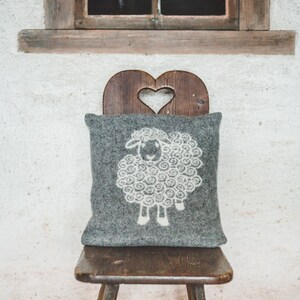 May include: A gray wool pillow with a white sheep design sits on a wooden chair with a heart-shaped cutout in the back. The chair is in front of a white wall with a wooden window above it.