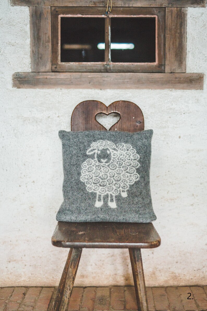 May include: A gray wool pillow with a white sheep design sits on a wooden chair with a heart-shaped cutout in the back. The chair is in front of a white wall with a wooden window above it.
