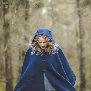 May include: A woman wearing a blue hooded cloak stands in a forest. The cloak has a button closure and is made of a soft, warm fabric.
