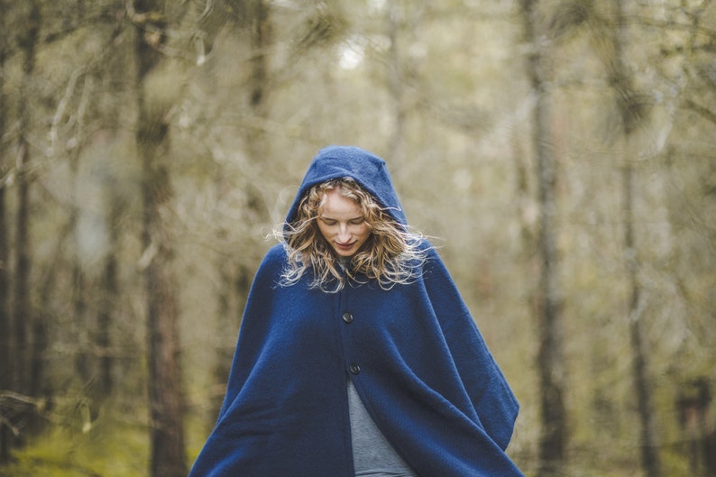 May include: A woman wearing a blue hooded cloak stands in a forest. The cloak has a button closure and is made of a soft, warm fabric.