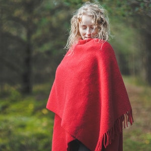 May include: A woman with long blonde curly hair is wearing a red shawl with fringe. She is standing in a forest with green moss on the ground.