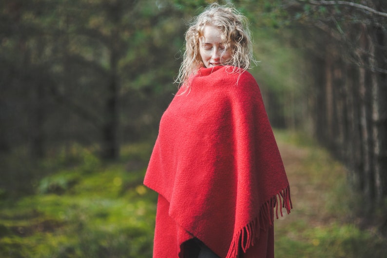 May include: A woman with long blonde curly hair is wearing a red shawl with fringe. She is standing in a forest with green moss on the ground.