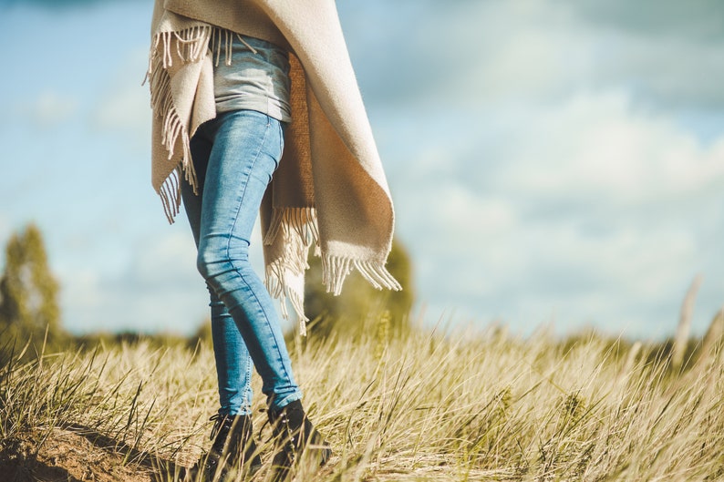 May include: A person wearing blue jeans and a beige fringed shawl walks through a field of tall grass.