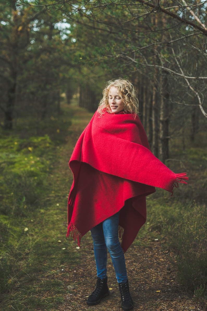 May include: A woman with curly blonde hair stands in a forest, wrapped in a red blanket. She is wearing blue jeans and black boots.