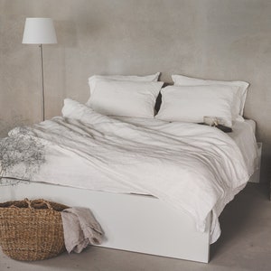 May include: A white linen duvet cover and pillowcases on a white bed frame with a wooden nightstand and a potted plant.