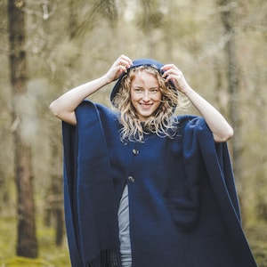 May include: A woman wearing a navy blue hooded poncho with a button closure. She is standing in a forest setting.