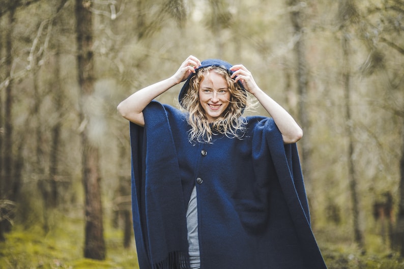 May include: A woman wearing a navy blue hooded poncho with a button closure. She is standing in a forest setting.