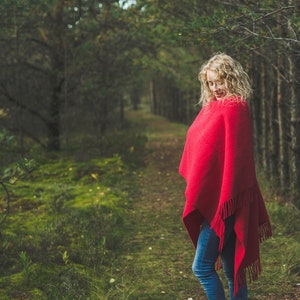 May include: A woman wearing a red shawl stands in a forest setting. The shawl has a fringe border and is draped over her shoulders. She is standing on a path in the woods, with green foliage and trees surrounding her.