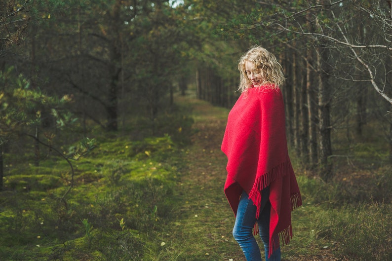 May include: A woman wearing a red shawl stands in a forest setting. The shawl has a fringe border and is draped over her shoulders. She is standing on a path in the woods, with green foliage and trees surrounding her.